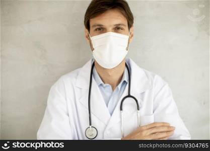 Attractive young male doctor wearing surgical protective mask and standing by the wall