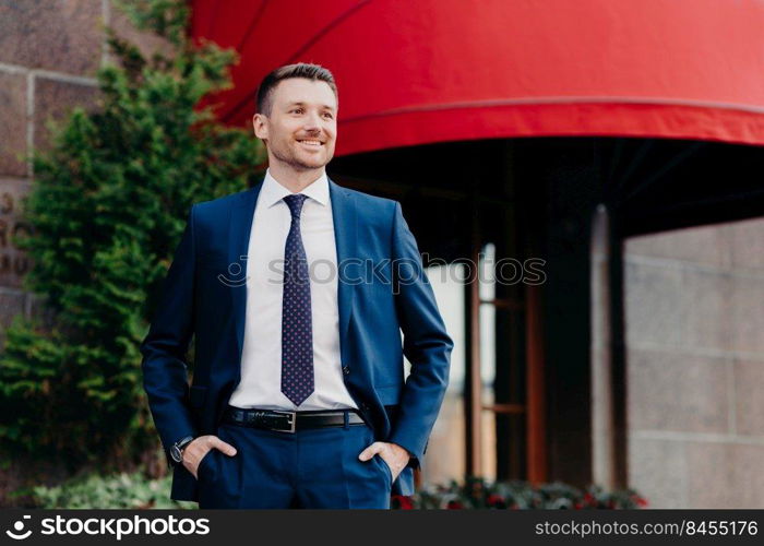 Attractive young male entrepreneur looks confidently and happily at camera, keeps hands in pockets, wears expensive suit, has positive smile, satisfied with achieving good results in business sphere