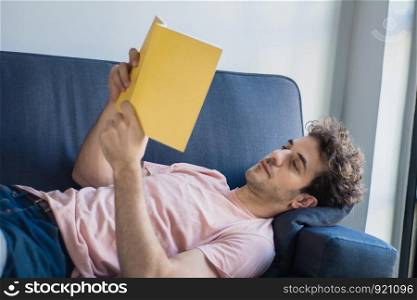 Attractive young man relaxed and reading a book on sofa at home. Indoors.