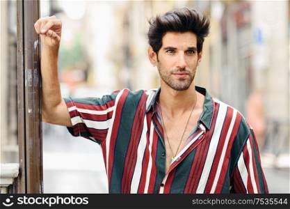 Attractive young man with dark hair and modern hairstyle wearing casual clothes in urban background. Guy with lost look.. Attractive young man with lost look wearing casual clothes outdoors