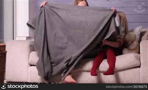 Attractive young mom warming her little daugther under the blanket while sitting on the couch together in the living room during cold winter evening. Mother hugging her cute girl with tenderness and love and wrapping her in blanket.