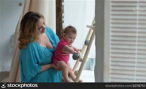 Attractive young mother holding her cute infant child, standing by the window at home. Adorable baby girl grasping decorative ball hanging on wooden stairs. Mom developing palmar grasp reflex of her little child.