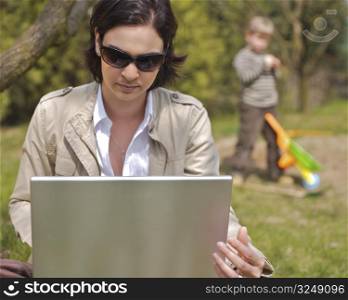 Attractive young mother is sitting on the ground in the garden and using a laptop while a 5 years old boy is playing in the background.