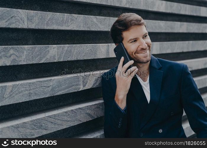 Attractive young office worker in stylish blazer speaking on mobile phone during work break, smiling sincerely while having nice family talk on smartphone while standing outside of business center. Attractive young man office worker in stylish blazer speaking on mobile phone outside