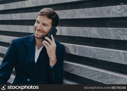 Attractive young office worker in stylish blazer speaking on mobile phone during work break, smiling sincerely while having nice family talk on smartphone while standing outside of business center. Attractive young man office worker in stylish blazer speaking on mobile phone outside