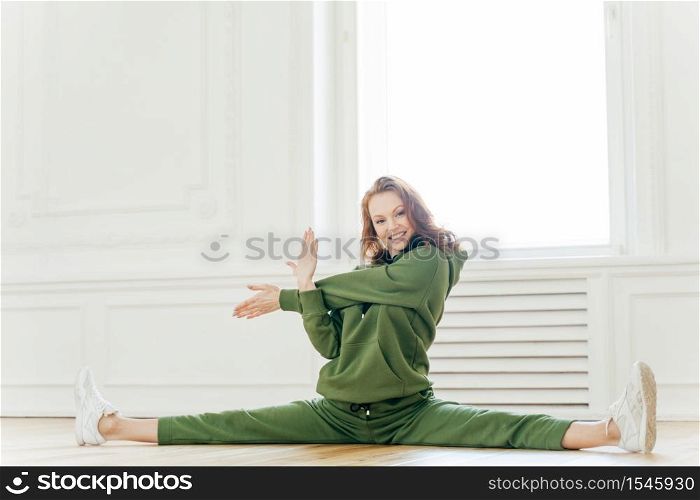 Attractive young slim woman makes splits, stretches hands, has flexible body, ginger wavy hair, dressed in sportswear, sits on floor, smiles at camera. People, training and healthy lifestyle concept