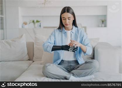 Attractive young woman adjusting her electronic artificial arm in living room. Handicapped european girl is changing settings of bionic arm. Cyber sensor hand has processor chip and buttons.. Attractive young woman adjusting her electronic artificial bionic arm in living room.