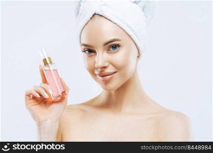 Attractive young woman applies parfum, enjoys pleasant scent, stands with naked shoulders, has natural makeup, healthy skin, wrapped towel on head after taking shower. Great aroma, try this.