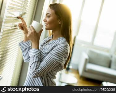 Attractive  young woman holding cup with hot tea or coffee and looking at the sunrise standing near the window in room