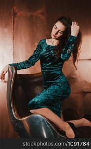 Attractive young woman in a blue green velvet dress, sitting in a leather brown chair, touching her long hair. Long legs, high heels. Luxury concept