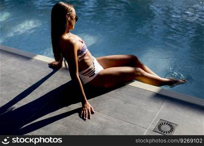 Attractive young woman in bikini and with sunglasses sits by the poolside and enjoys the summer sun