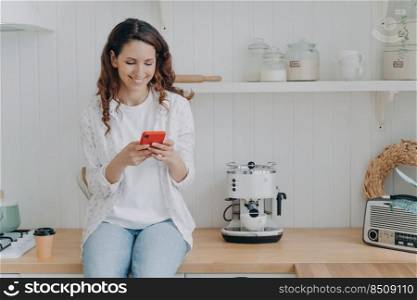 Attractive young woman is texting on smartphone. Housewife has leisure and sitting on worktop at kitchen at home. Smiling girl with telephone. Modern luxurious scandinavian interior.. Attractive young woman is texting on smartphone. Girl sitting on worktop at kitchen at home.