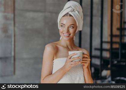 Attractive young woman puts moisturizing cream on face after showering, smiles broadly, wrapped in towels, drinks coffee, poses indoor, thinks about something pleasant. Beauty skin care concept
