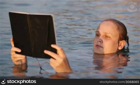 Attractive young woman reading a book while floating on her back in a tranquil sea