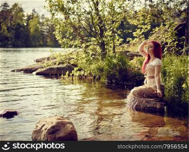 Attractive young woman sits on the rock and she dreams, summer day on the lake
