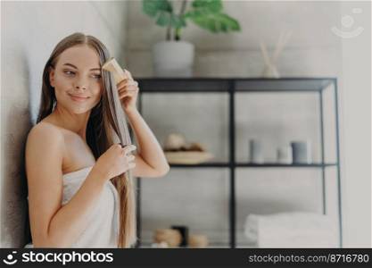 Attractive young woman smiles and enjoys her beauty, combes long straight hair after applying mask, stands wrapped in bath towel in cozy bathroom, has smooth skin. Hair care, spa and beauty concept