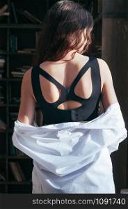 Attractive young woman wearing a black sport bra lingerie, white shirt disclosing shoulders, back view. Casual fashion style, posing by the window. Sensual beautiful seductive. For catalogue