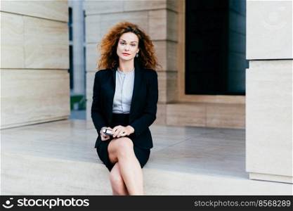Attractive young woman wearing black formal suit, sitting crossed legs with electronic device waiting for business partner near cafe. Gorgeous female with appealing appearance posing at camera