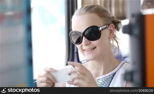 Attractive young woman wearing sunglasses holding up and photographing at her mobile on a bus or train