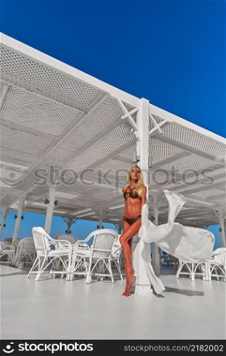 Attractive young woman wearing swimsuit and pareo standing over white wooden wall.. Attractive young woman wearing swimsuit and pareo standing over white wooden wall