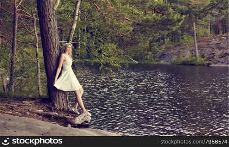 Attractive young woman wearing white summer dress and she leans towards the pine, sunny summer day, lake on background
