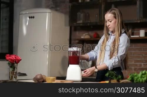Attractive young woman with amazing blonde long hair making beet smoothie with blender in grunge rustic styled kitchen. Beautiful female preparing healthy organic beet smoothie at home.