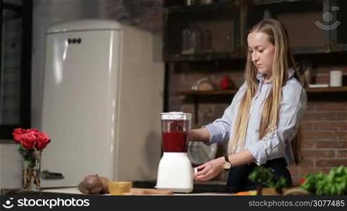 Attractive young woman with amazing blonde long hair making beet smoothie with blender in grunge rustic styled kitchen. Beautiful female preparing healthy organic beet smoothie at home.