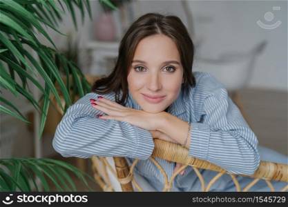 Attractive young woman with dark hair, nice make up looks directly at camera, leans on back of wicker furniture, domestic atmosphere, green plant, has confident expression. People and rest concept