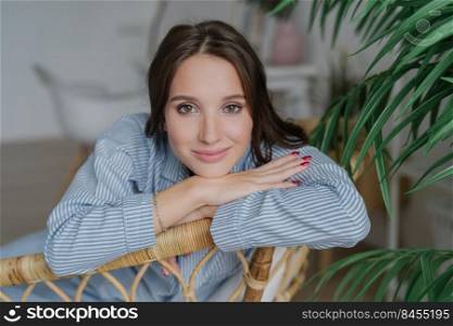 Attractive young woman with dark hair, nice make up looks directly at camera, leans on back of wicker furniture, domestic atmosphere, green plant, has confident expression. People and rest concept