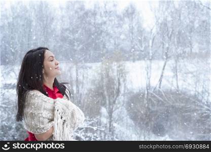 Attractive young woman with long dark hair, in a red dress, covered by a handmade shawl, with her eyes closed, enjoying the snowfall.