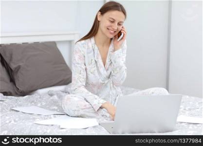 Attractive Young Woman Working from Home - Female Entrepreneur Sitting on Bed with Laptop Computer, Paperwork and talking on the Cell Phone from Comfort of Home. quarantine. coronavirus.. Attractive Young Woman Working from Home - Female Entrepreneur Sitting on Bed with Laptop Computer, Paperwork and talking on the Cell Phone from Comfort of Home. quarantine. coronavirus