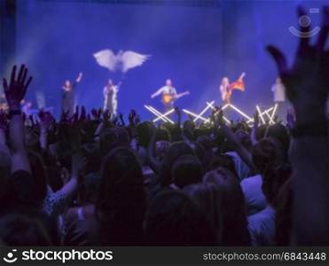 Audience with hands raised at a music festival and lights streaming down from above the stage. Focus on hands