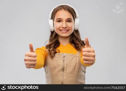 audio equipment and technology people concept - smiling teenage girl in headphones listening to music and showing thumbs up over grey background. teenage girl in headphones showing thumbs up