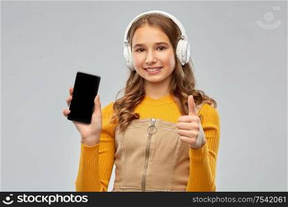 audio equipment and technology people concept - smiling teenage girl in headphones listening to music on smartphone and showing thumbs up over grey background. teenage girl in headphones showing smartphone
