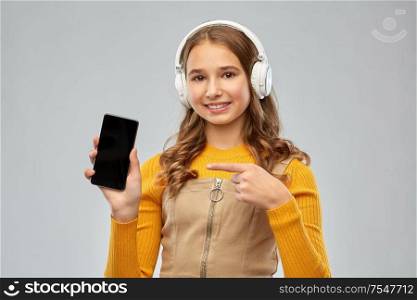 audio equipment and technology people concept - smiling teenage girl in headphones listening to music and showing smartphone over grey background. teenage girl in headphones showing smartphone