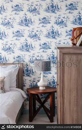 AUG 4, 2014 Hua Hin, THAILAND - Vintage cozy hotel room with bed, white ceramic lamp, pillows, blue pattern wallpaper, wooden side table and cabinet. Vintage country home decoration