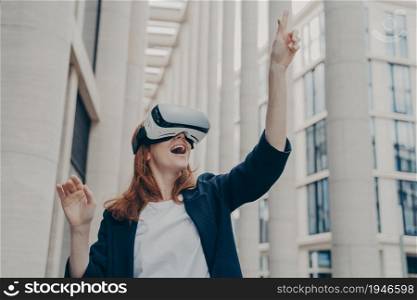 Augmented reality in business. Young excited happy female enterpruer wearing portable VR goggles trying to touch something in virtual reality with her finger, standing alone on city street. Excited female enterpruer wearing portable VR goggles trying to touch something in virtual reality