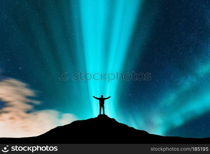 Aurora and silhouette of standing man with raised up arms on the mountain. Lofoten islands, Norway. Aurora borealis and happy man. Sky with stars and green polar lights. Night landscape with traveler