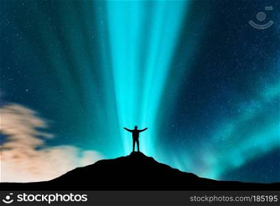 Aurora and silhouette of standing man with raised up arms on the mountain. Lofoten islands, Norway. Aurora borealis and happy man. Sky with stars and green polar lights. Night landscape with traveler