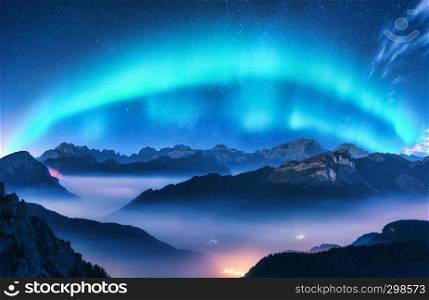 Aurora borealis above mountains in fog at night. Northern lights. Sky with stars with polar lights and high rocks. Beautiful landscape with aurora, city lights in low clouds, mountain ridge. Space