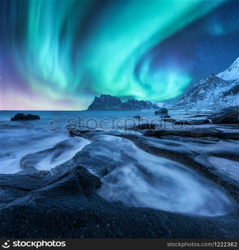 Aurora borealis above snowy mountains and sandy beach with stones. Northern lights in Lofoten islands, Norway. Starry sky with polar lights. Night winter landscape with aurora, sea with blurred water