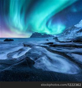 Aurora borealis above snowy mountains and sandy beach with stones. Northern lights in Lofoten islands, Norway. Starry sky with polar lights. Night winter landscape with aurora, sea with blurred water