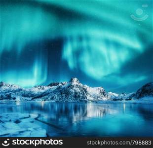 Aurora borealis above snowy mountains, frozen sea coast and reflection in water in Lofoten islands, Norway. Northern lights. Winter landscape with polar lights, ice in water. Sky with stars and aurora. Aurora borealis above snowy mountains, frozen sea coast
