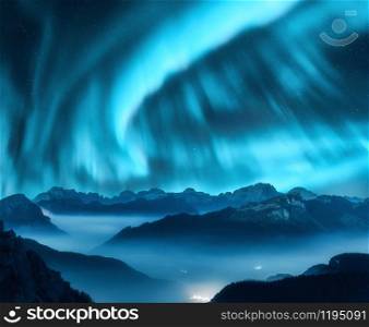 Aurora borealis above the mountains in fog at night. Northern lights. Sky with stars with polar lights and high rocks. Beautiful landscape with aurora, city lights in low clouds, mountain peaks. Space