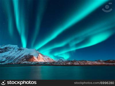 Aurora borealis above the snow covered mountain in Lofoten islands, Norway. Northern lights in winter. Night landscape with polar lights, snowy rocks, reflection in the sea. Starry sky with aurora