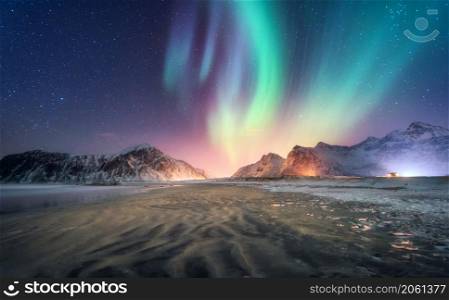 Aurora borealis above the snowy mountain and sandy beach in winter. Northern lights in Lofoten islands, Norway. Starry sky with polar lights. Night landscape with aurora, frozen sea coast, city lights