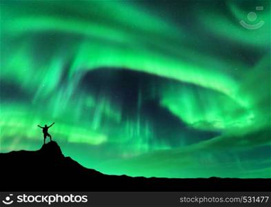 Aurora borealis and silhouette of a man with raised up arms on the mountain peak. Lofoten islands, Norway. Aurora and happy man. Sky with stars and polar lights. Night landscape with aurora and people