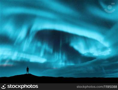 Aurora borealis and silhouette of standing man on the hill. Lofoten islands, Norway. Aurora and alone man. Blue sky with stars and bright polar lights. Night landscape with aurora and people. Travel
