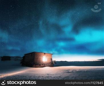 Aurora borealis over old small house with light from the window in winter. Northern lights in Teriberka, Russia. Blue starry sky and green polar lights. Night landscape with aurora and building