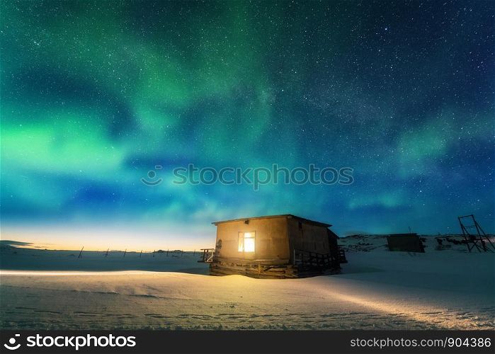 Aurora borealis over old small house with yellow light from the window. Northern lights in Teriberka, Russia. Blue sky with stars and green polar lights. Night winter landscape with aurora, building